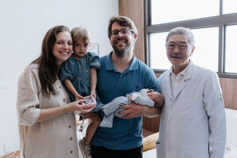 Retrato oficial da família reunida no quarto da maternidade ao lado do médico obstetra sorridente.'