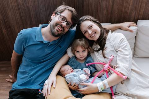 Retrato oficial da família de quatro pessoas reunida e sorridente no sofá do quarto do hospital.'
