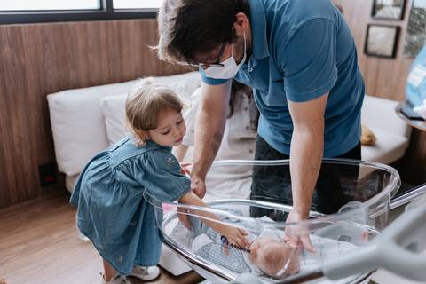 Foto do pai e da filha inclinados sobre o berço móvel de acrílico, fazendo carinho no bebê Pedro.'