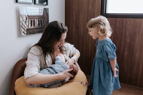 Foto da mãe sentada na poltrona amamentando o bebê Pedro com o apoio da almofada amarela, enquanto a filha mais velha observa de perto.'