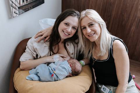 Retrato da avó sorridente abraçada à mãe, que está sentada na poltrona amamentando o pequeno Pedro no quarto do hospital.'