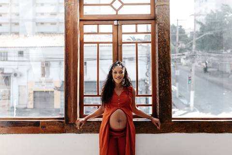 Gestante posando sorridente em frente a uma grande janela de madeira com vista para a cidade.'