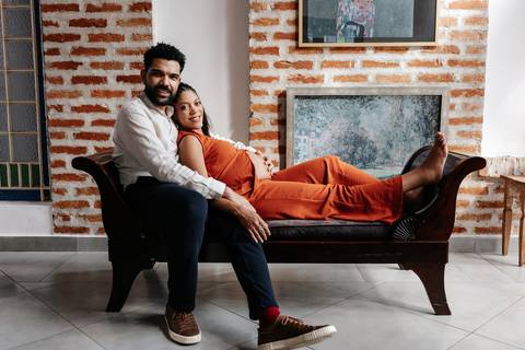 Retrato do casal relaxado em um recamier de madeira, com a gestante deitada de costas apoiada no parceiro.'