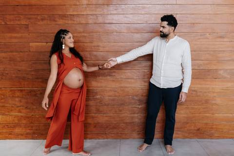 Foto de ângulo aberto do casal em pé de mãos dadas, com distanciamento poético diante da parede de madeira.'