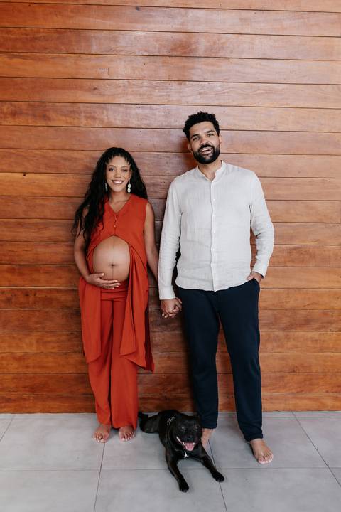 Retrato do casal posando lado a lado de mãos dadas, destacando a barriga da gestante em primeiro plano.'