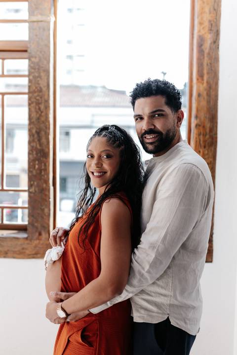 Retrato do casal sorridente em perfil de frente para a janela, com o parceiro abraçando a gestante por trás.'