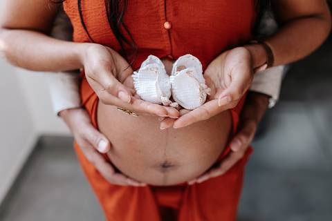Close focado nas mãos do casal segurando um par de sapatinhos brancos de bebê logo acima da barriga exposta.'
