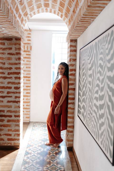 Retrato da gestante em pé entre pilares de tijolos aparentes, destacando sua silhueta de perfil.'