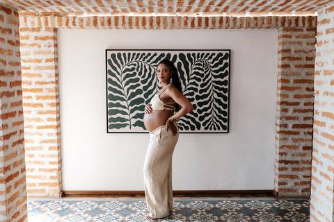 Foto de ângulo aberto da gestante com um top e calça clara, posando de perfil em um corredor com piso desenhado e quadro de folhagem ao fundo.'