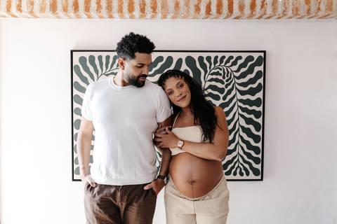 Retrato do casal abraçado com roupas claras e casuais, posando em frente ao quadro de folhagem em preto e branco.'