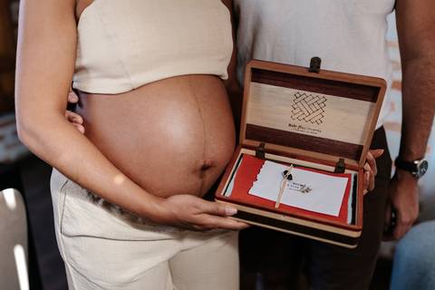 Detalhe focado nas mãos do casal segurando uma caixinha de madeira aberta com recordações ao lado da barriga.'