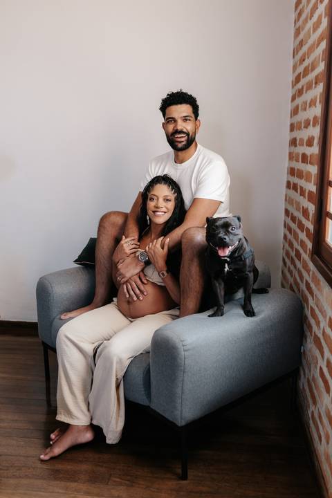 Retrato do casal sentado no braço do sofá cinza com o seu cachorro de estimação preto posando ao lado.'
