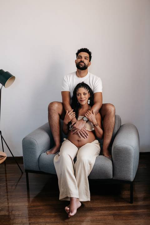 Foto centralizada do casal posando no sofá; o parceiro está posicionado por trás abraçando a barriga da gestante.'