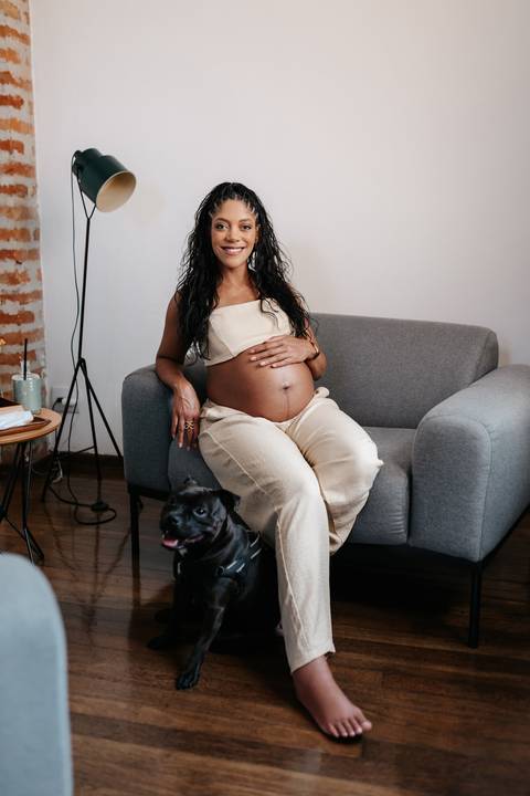 Retrato da gestante sentada de forma descontraída no sofá cinza com a barriga à mostra e o cão em primeiro plano.'