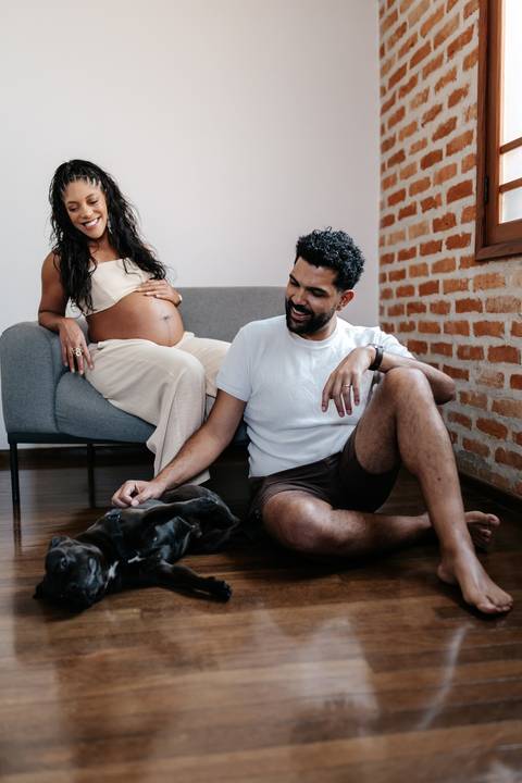 Registro espontâneo do casal rindo no chão da sala com o cachorro de estimação relaxado entre eles.'