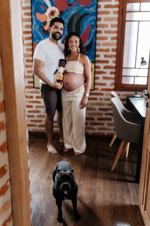 Foto de ângulo aberto mostrando o casal em pé na sala com tijolos aparentes, destacando a calmaria do ambiente.'