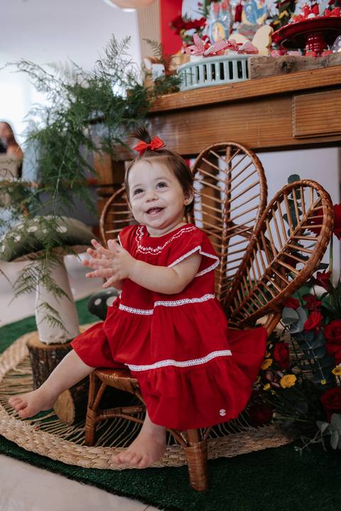 Aniversariante de vestido vermelho batendo palminhas na cadeira de vime.'