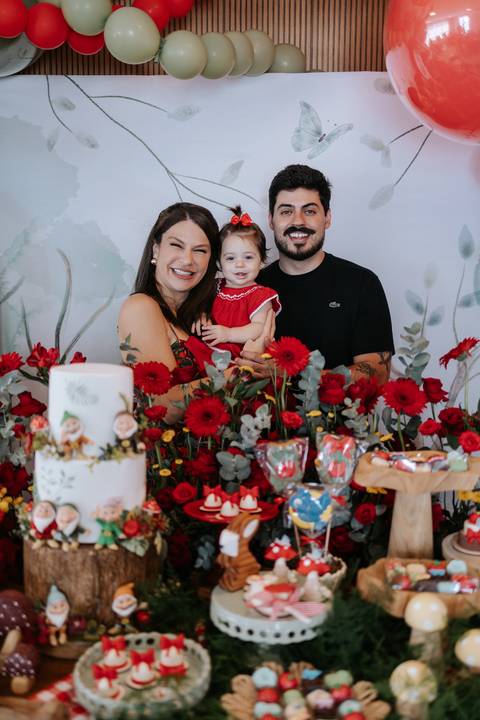 Pais com a filha no colo atrás da mesa cheia de flores vermelhas.'