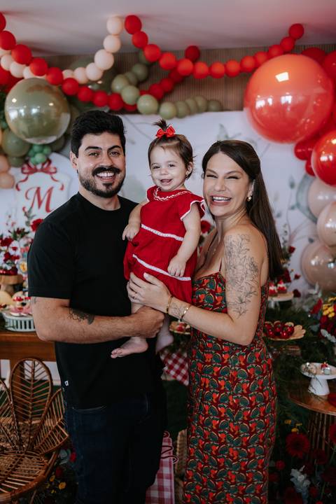 Retrato do casal com a bebê em frente ao painel de balões.'