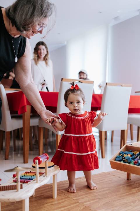 Aniversariante de vestido vermelho em pé no salão segurando a mão de um adulto.'