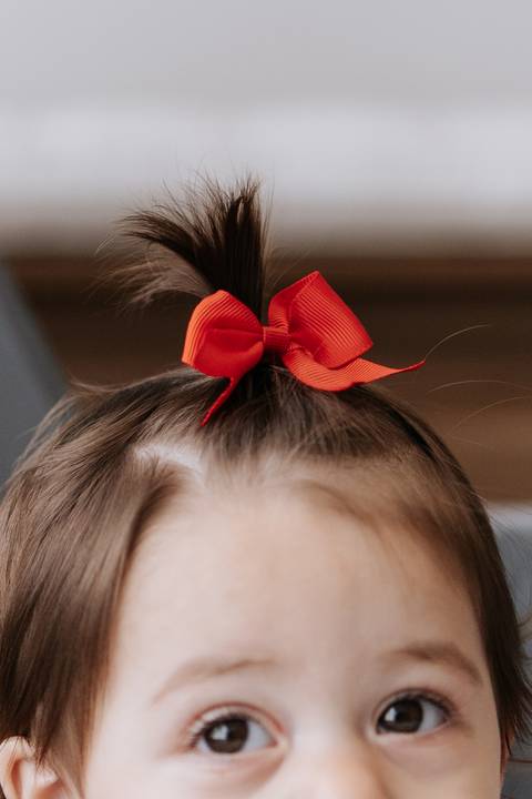 Detalhe em close do laço vermelho de fita no cabelo da bebê.'