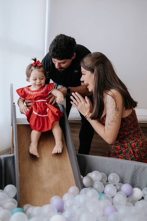 Pais sorrindo e aplaudindo a filha no topo do mini escorregador.'