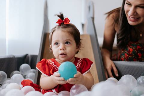 Aniversariante concentrada segurando uma bolinha azul enquanto a mãe observa ao fundo.'