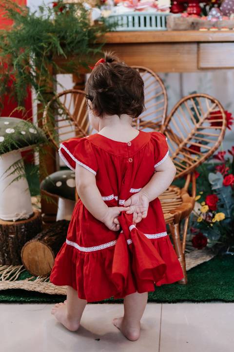 Detalhe das costas do vestido vermelho da bebê enquanto ela caminha pelo cenário.'