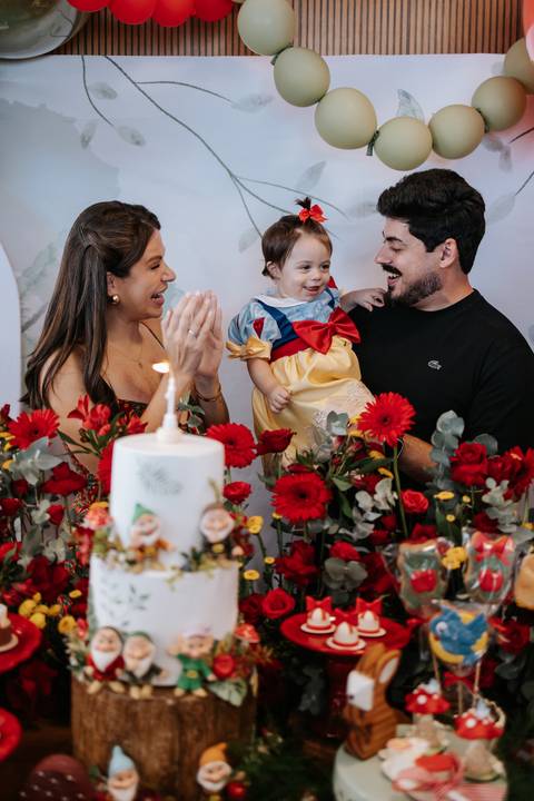 Pais sorrindo e batendo palmas com a aniversariante vestida de Branca de Neve diante do bolo.'