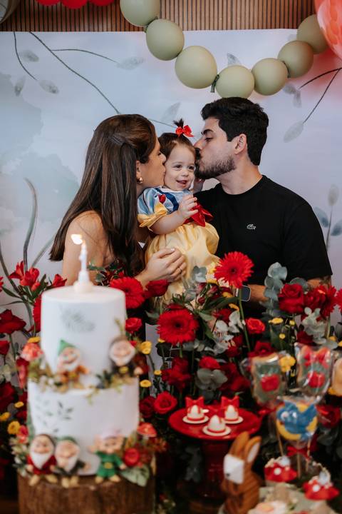 Pais dando beijinhos na filha com a vela do bolo acesa em primeiro plano.'