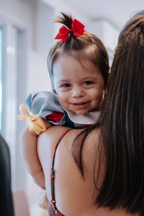 Close fofinho da aniversariante sorrindo por cima do ombro da mãe.'