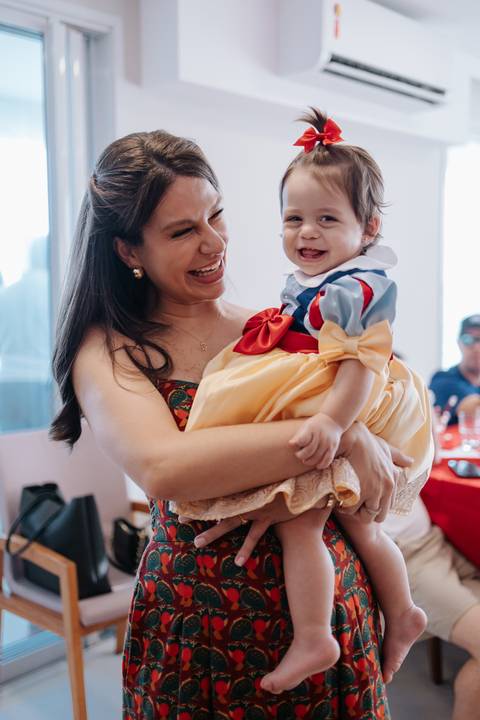 Mãe sorrindo e segurando a aniversariante no colo em momento descontraído.'