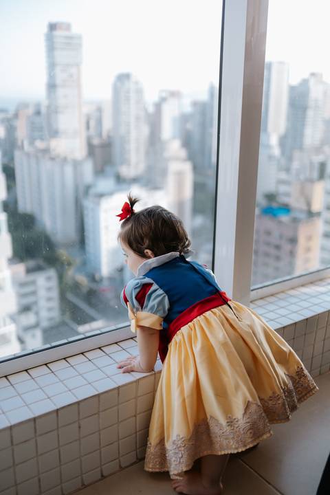 Bebê de costas vestida de Branca de Neve, olhando pela grande janela de vidro do salão.'