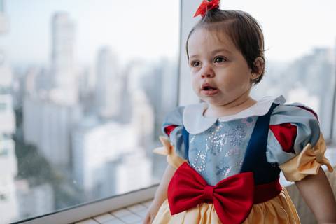 Close-up da aniversariante com sua fantasia de princesa, posando em frente à janela com vista da cidade ao fundo.'
