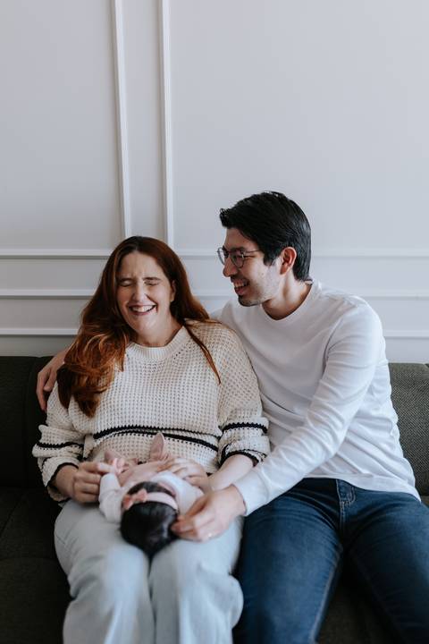 Casal sorridente posando com o bebê nos braços no ambiente da sala.'