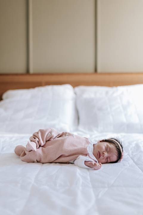 ebê de macacão rosa deitada de costas na grande cama de casal branca.'