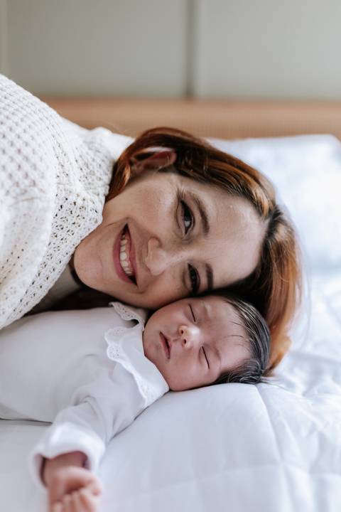 Close da mãe sorridente com o rosto colado ao da bebê deitada.'