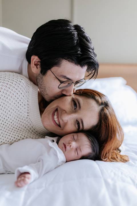 Retrato do casal sorrindo com os rostos bem pertinho da bebê que dorme na cama.'
