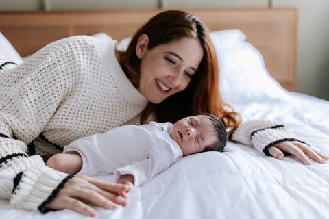 Mãe deitada na cama observando a bebê recém-nascida dormir ao seu lado.'