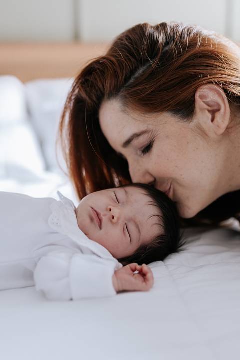 Mãe dando um beijo terno na testa da bebê que dorme na cama.'