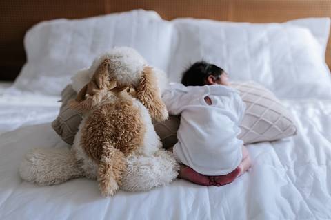 Recém-nascida deitada de bruços na cama ao lado de um grande bicho de pelúcia.'
