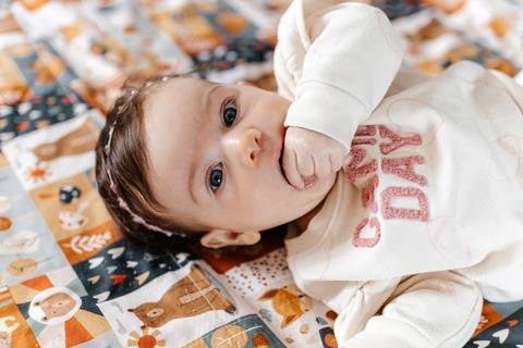 Close da bebê deitada de costas sobre um tapetinho estampado colocando a mão na boca.'