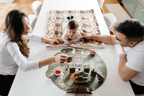Visão aérea dos pais sentados à mesa com a bebê deitada em cima do tapete decorativo.'