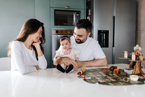 Pais olhando e sorrindo para a bebê sentada no colo no ambiente da cozinha.'