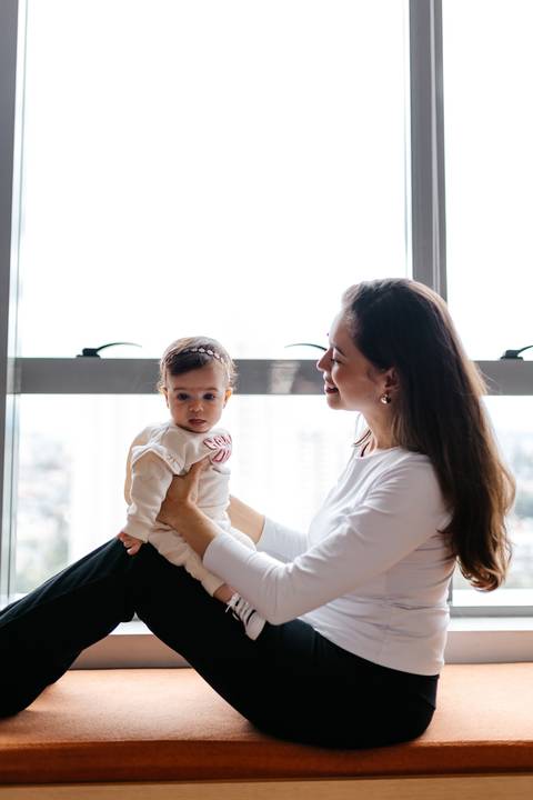 Mãe segurando a bebê no colo em frente à grande janela iluminada.'