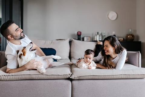 Pais e a bebê sentados no sofá com o cachorro deitado confortavelmente ao lado.'
