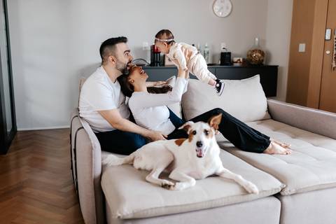 Retrato do casal no sofá erguendo a bebê no ar com o cachorro deitado ao lado.'