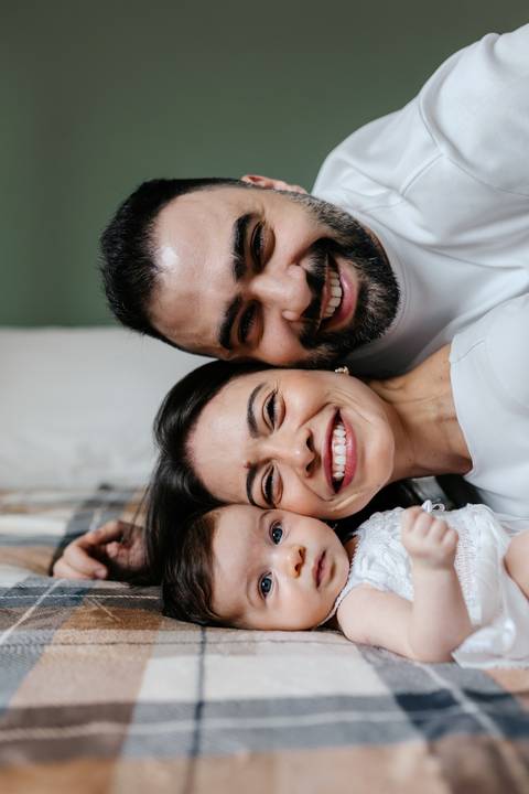 Close do casal deitado na cama sorrindo de cabeça para baixo com a bebê.'