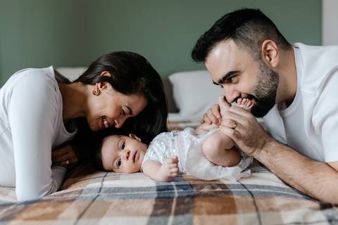 Pais deitados na cama interagindo e brincando com a bebê entre eles.'
