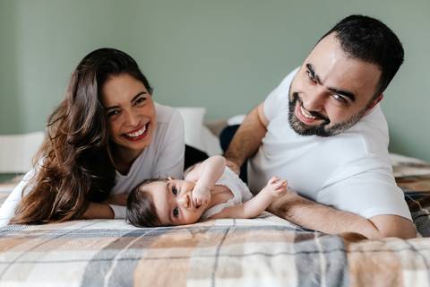 Casal de bruços na cama sorrindo e se divertindo com a bebê no centro.'